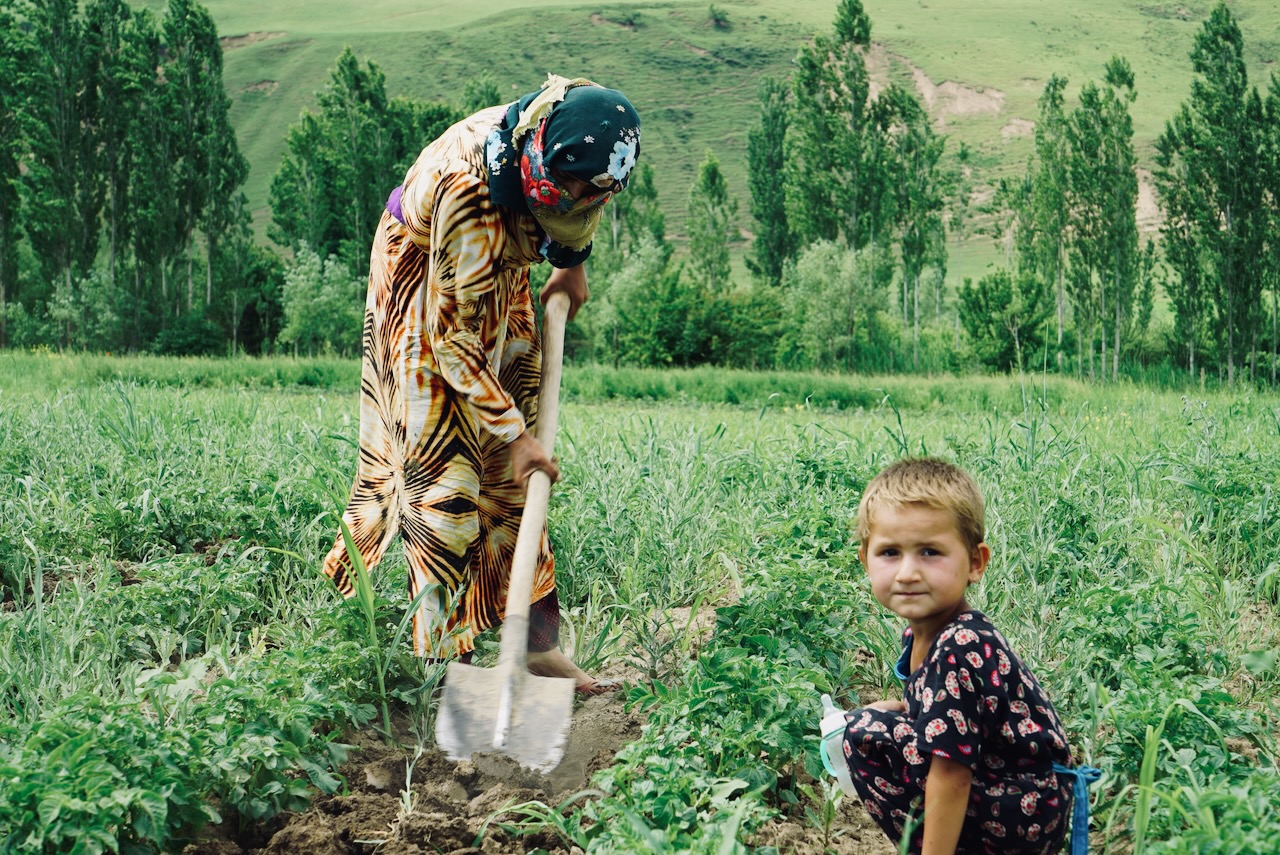A woman works the soil with a shovel while a child crouches beside her