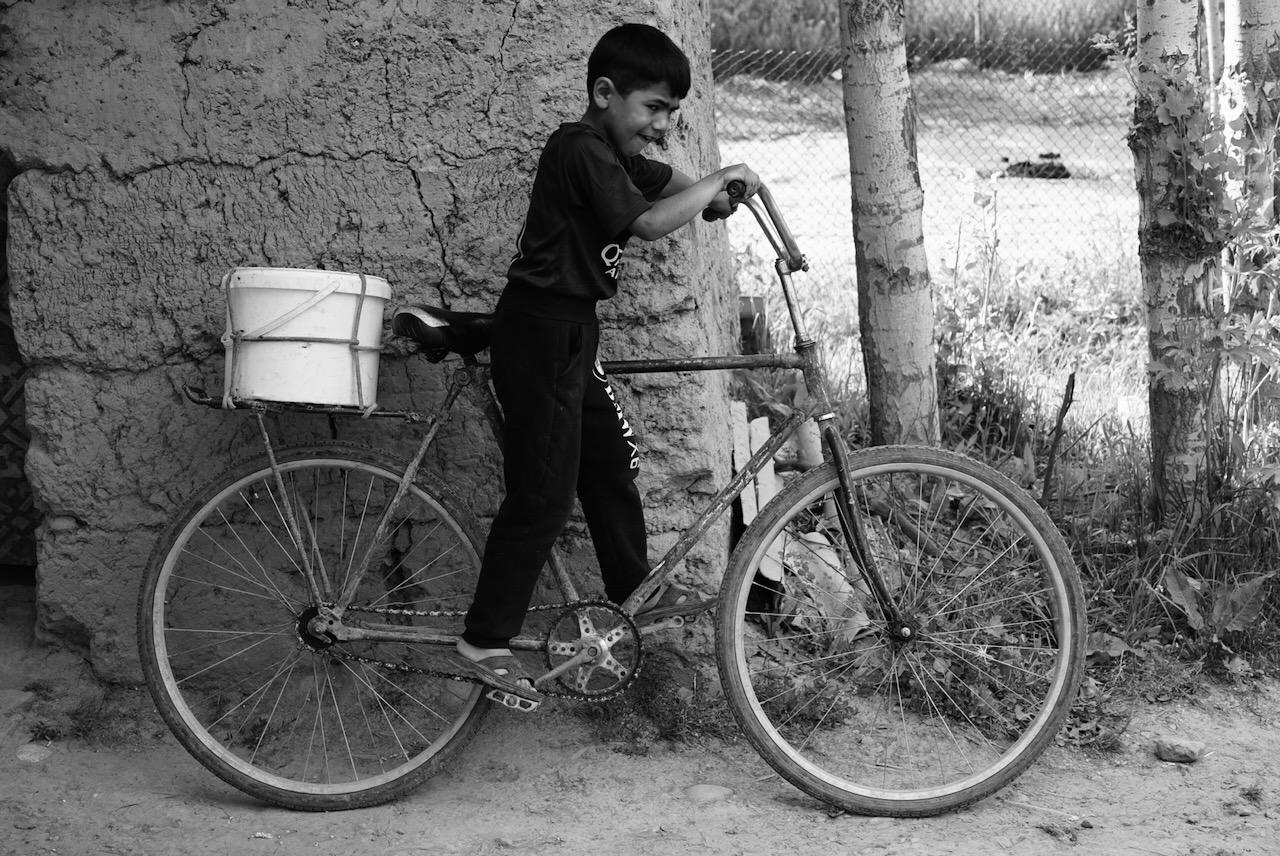 A boy with a bucket strapped to a bicycle, leaning against an adobe wall