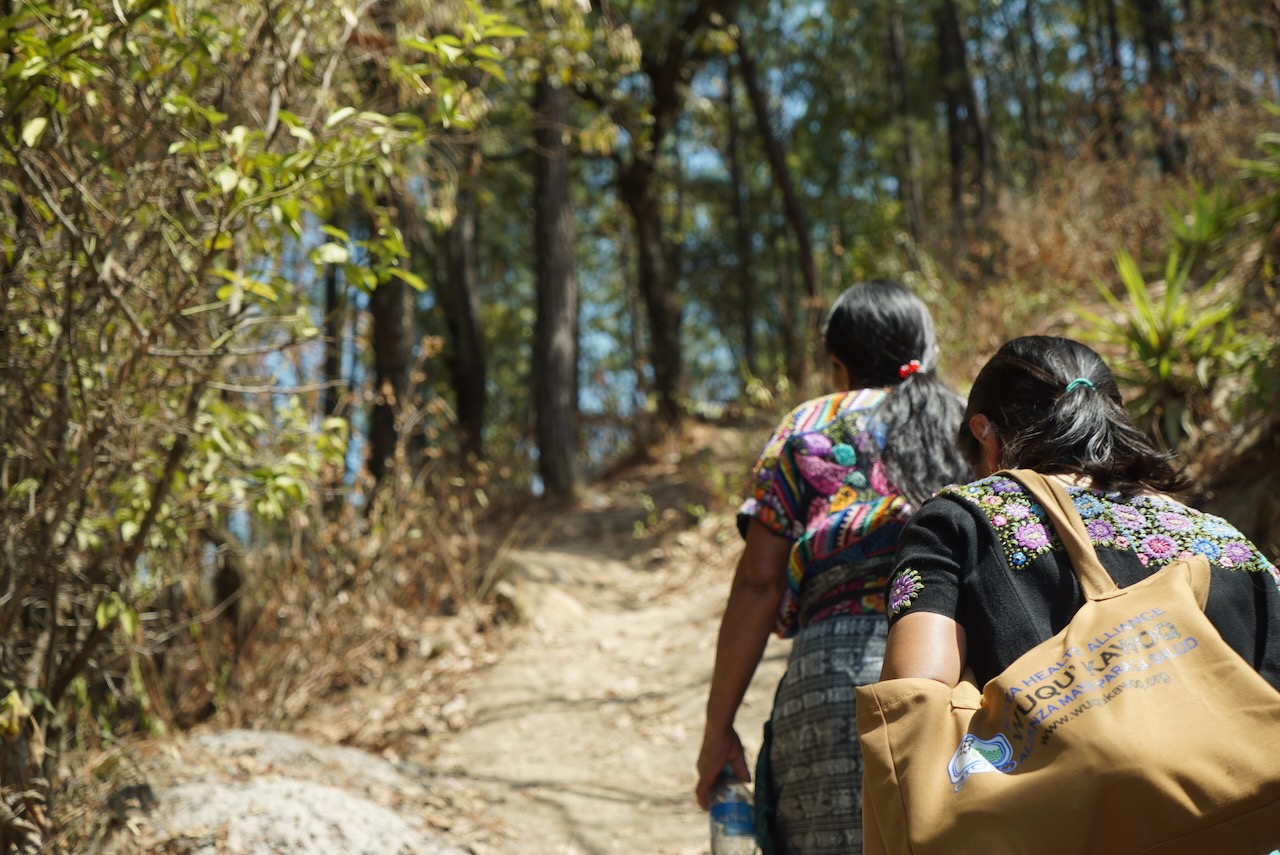 Two health workers walking a forest trail, one carrying a Maya Health Alliance bag
