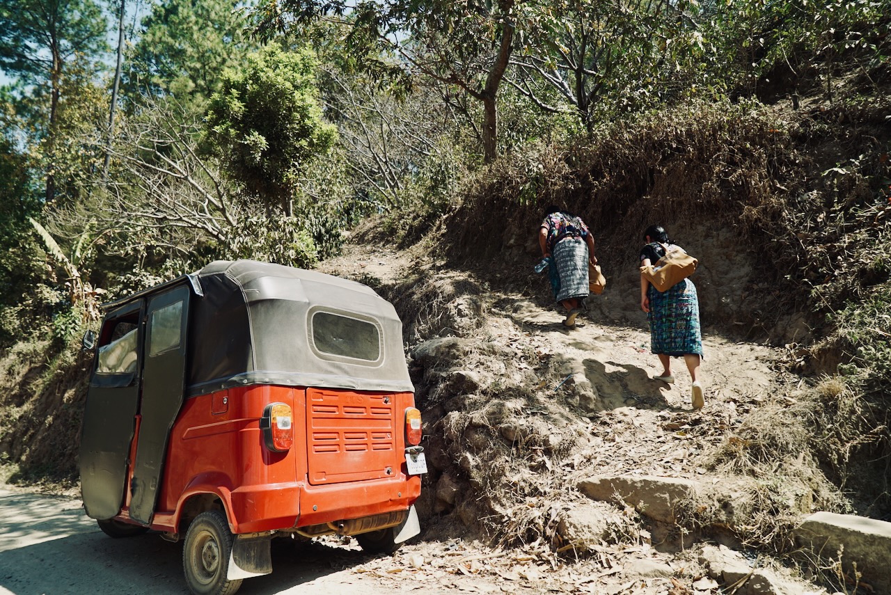 Two health workers climb a dirt path past a red tuk-tuk in the Guatemalan highlands